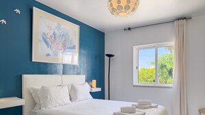 Uma cama bem arrumada com lençóis brancos, uma mesa de cabeceira com abajur, um quadro abstrato na parede e uma janela com cortinas.