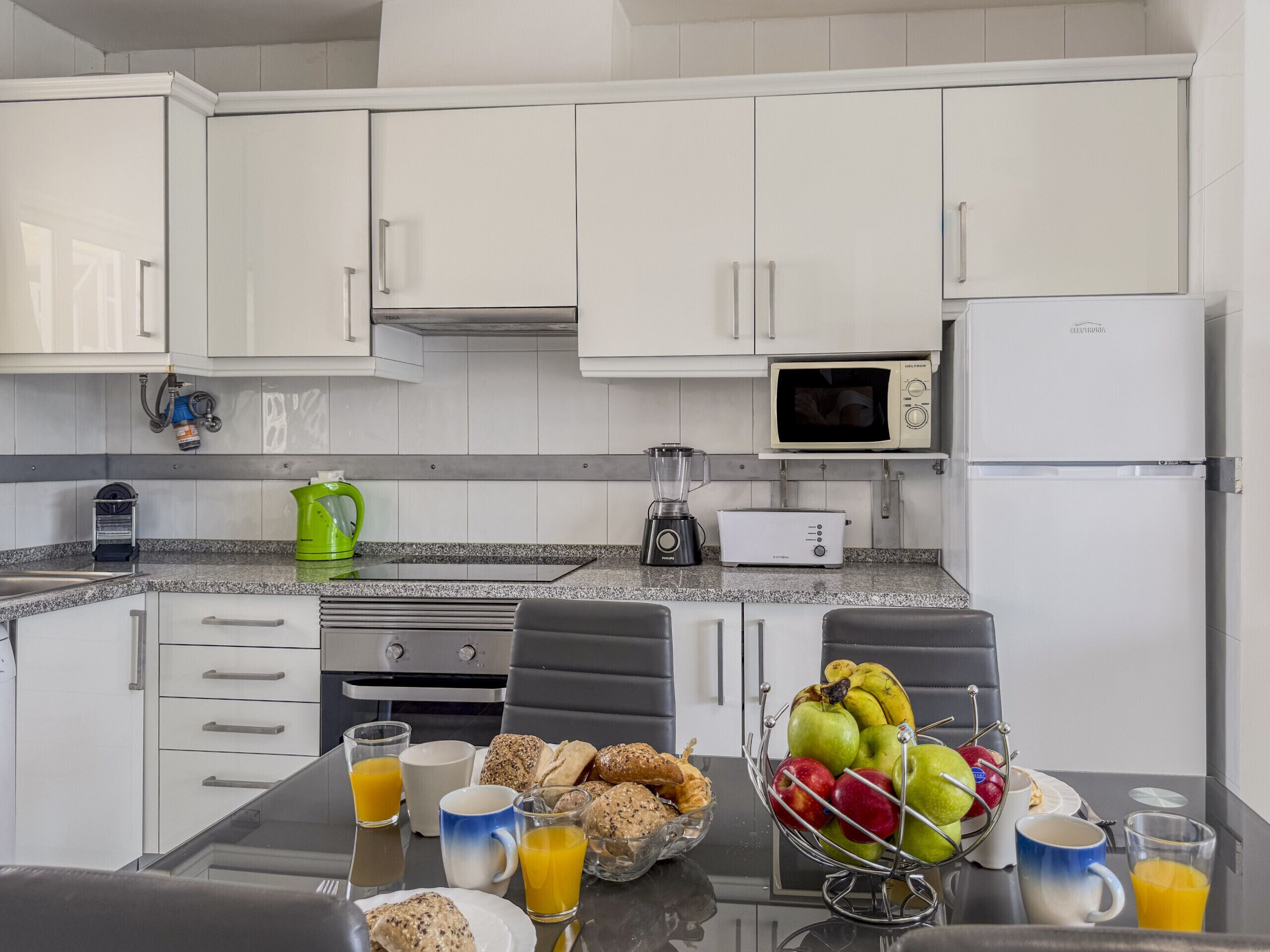 Uma cozinha moderna com armários brancos, micro-ondas, frigorífico, máquina de café e uma mesa de jantar posta para o pequeno-almoço.