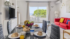Uma sala de jantar com mesa posta para o café da manhã, um sofá vermelho e uma varanda com vista para o mar.