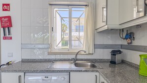 Uma cozinha com pia de aço inoxidável, uma janela com cortina, uma máquina de lavar louça e uma chaleira verde.
