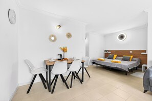 Ein modernes, helles Zimmer mit Essbereich, einem Bett mit Holz-Kopfteil und einem kleinen Tisch mit einer Vase.