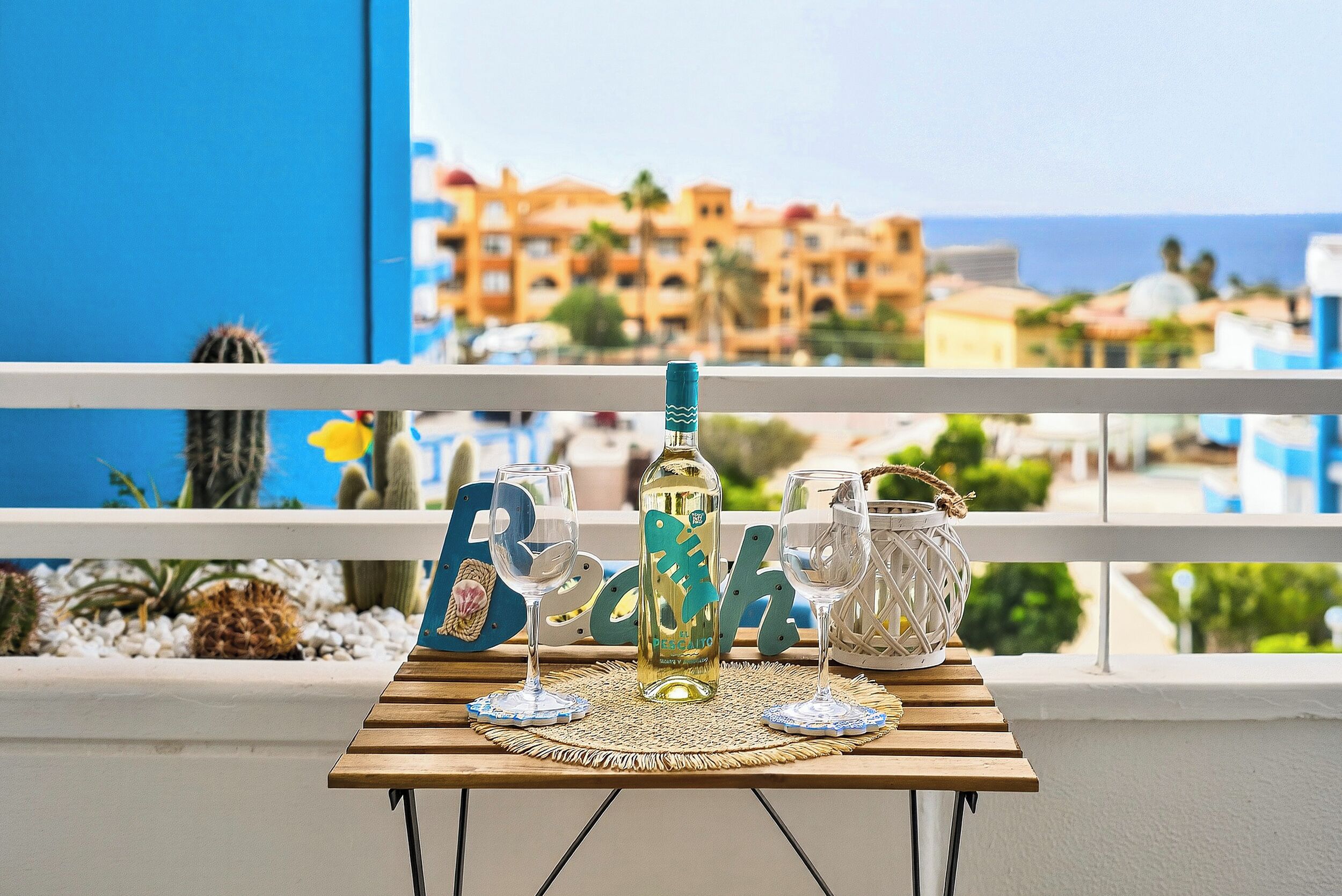 Ett bord med en flaska, glas och dekorativa föremål på en balkong med utsikt över ett kustnära område, där byggnader syns i bakgrunden.