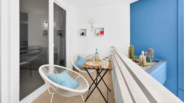 En balkong med flätade stolar, ett litet bord med en flaska, glas och en dekorativ detalj. Det finns kaktusar i en kruka och en blå vägg.