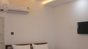 Un dormitori amb un llit de fusta, roba de llit blanca, una manta vermella, un ventilador de sostre, un aparell d'aire condicionat i una televisió penjada.