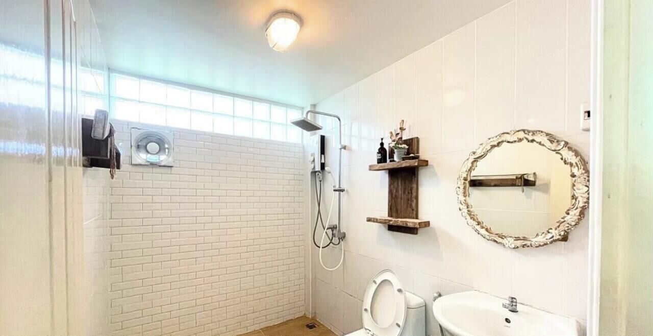 Baño con ducha, inodoro y lavamanos, con paredes de ladrillo blanco y un estante de madera con artículos de aseo.