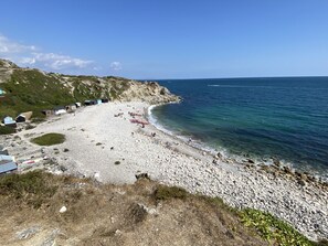 A pebble beach with a rocky shoreline, a clear blue sky, and a few buildings in the background.