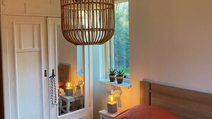 Une chambre à coucher avec un lit, un luminaire suspendu, une armoire blanche et une fenêtre ornée de plantes.