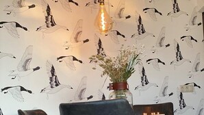 Un coin repas comprenant une table, deux chaises, un luminaire suspendu et un mur orné d’un motif représentant des oiseaux.