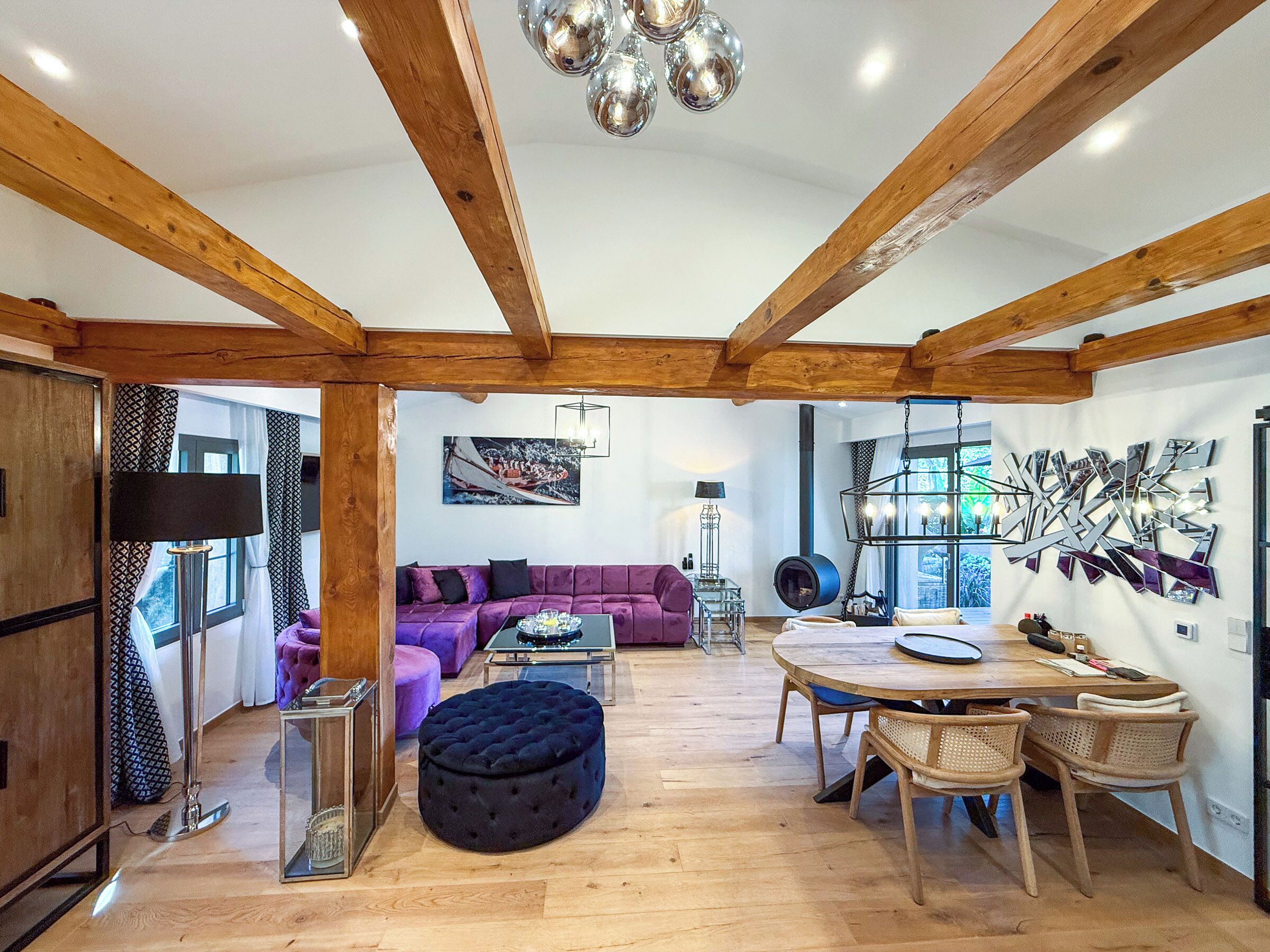 A spacious room with wooden beams, a purple sofa, a round table, and a hanging light fixture.