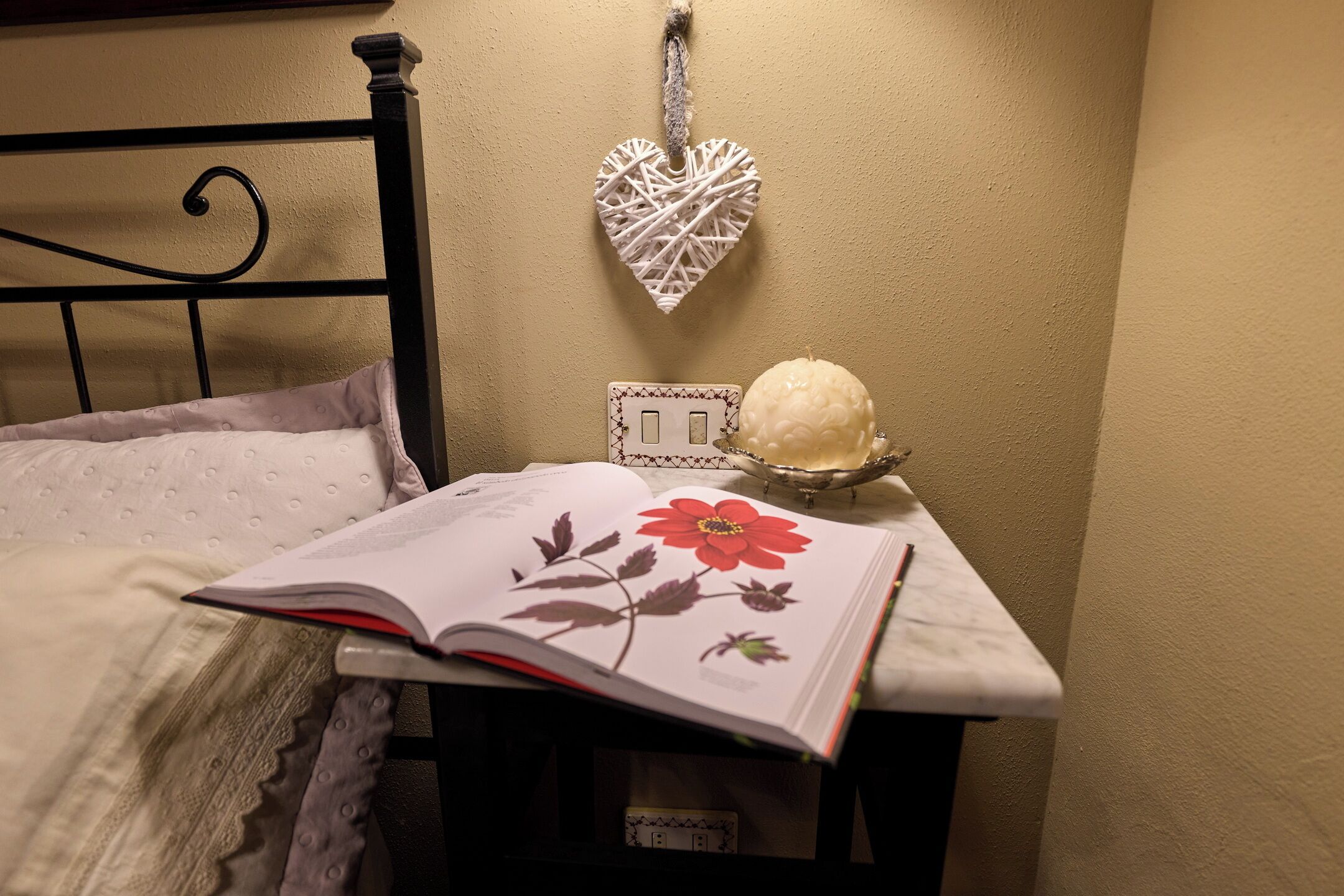 Un lit avec un oreiller blanc, un livre illustré d’une fleur rouge, un cœur décoratif et un petit objet blanc posé sur une table.