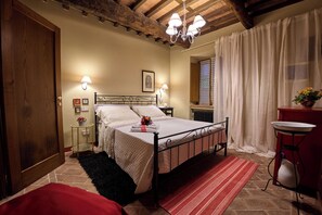 Ein Schlafzimmer mit einem Bett, Nachttischlampen, einem roten Teppich, einer Holzbalkendecke, einem Fenster mit Vorhängen und einem kleinen Tisch mit Blumen in einer Vase.