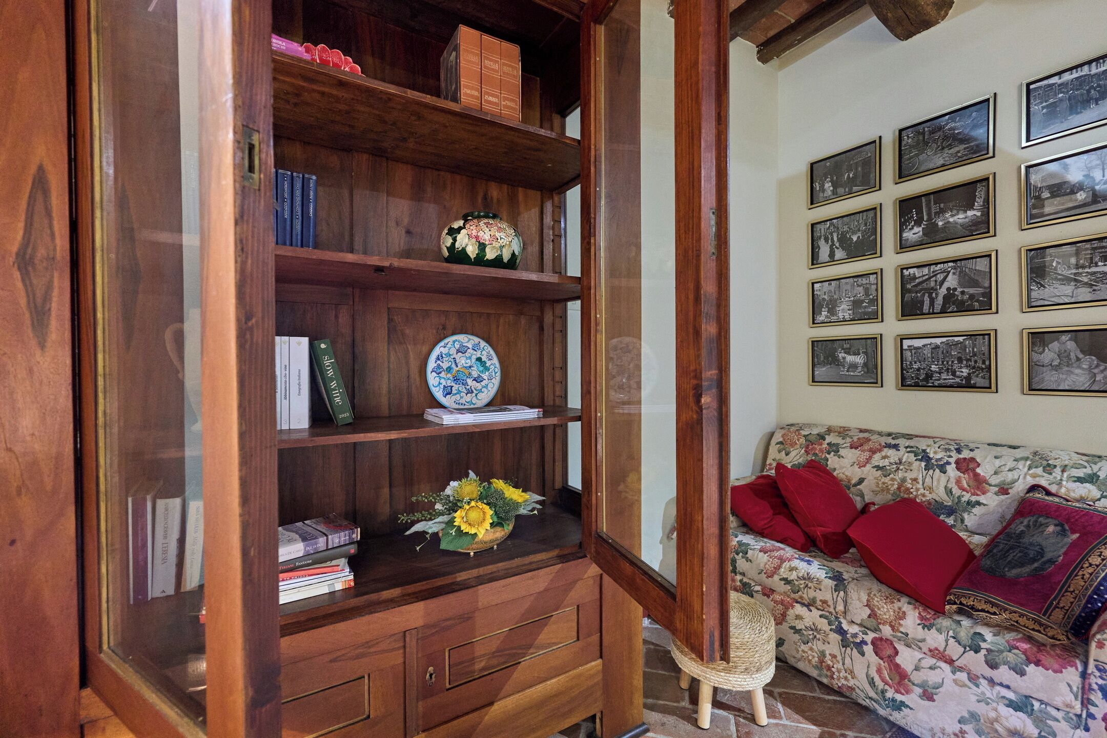 Een houten boekenkast met boeken en decoratieve objecten, een bloemenbank met rode kussens, en een wand met ingelijste foto's.