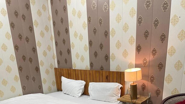 A hotel room with a wooden bed, a bedside table with a lamp, a chair, and patterned wallpaper.