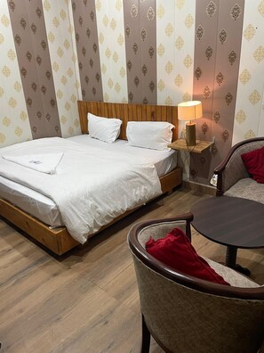 Una habitación de hotel con una cama de madera, una mesita de noche con lámpara, una silla y papel tapiz estampado.