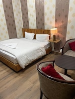 A hotel room with a wooden bed, a bedside table with a lamp, a chair, and patterned wallpaper.