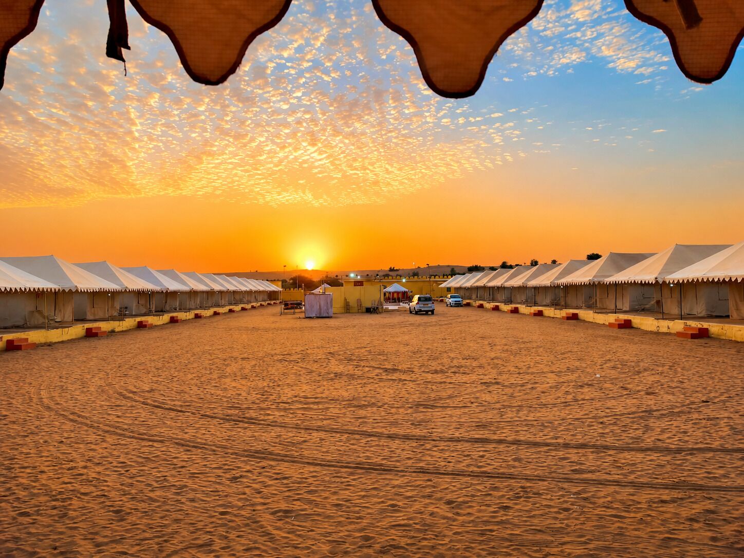 A desert camp with multiple tents arranged in rows, a clear sky, and the sun setting in the background.