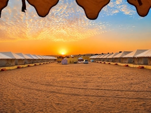 A desert camp with multiple tents arranged in rows, a clear sky, and the sun setting in the background. - Melody Moon Desert Camp (Sam)
