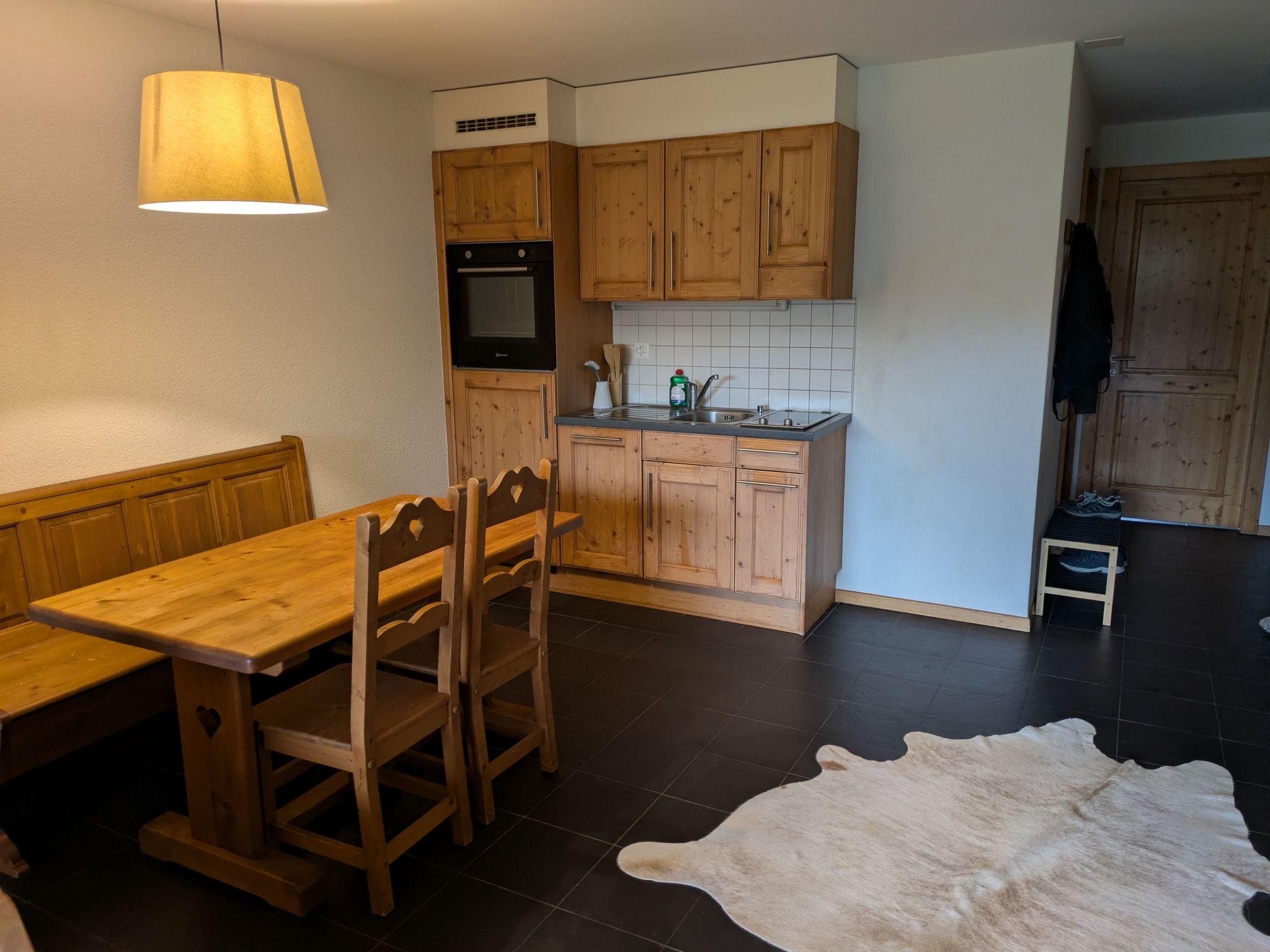 Une cuisine avec des meubles de rangement en bois, une table à manger avec des chaises, un luminaire suspendu et un tapis sur le sol.