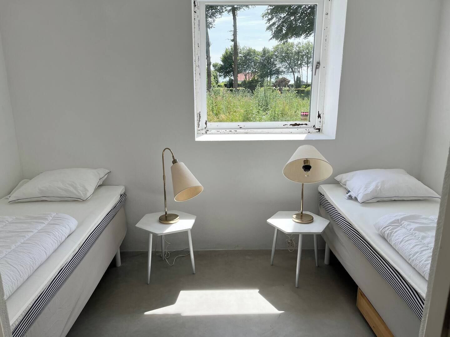 A small, bright room with two single beds, a window with a view of greenery, and two bedside lamps.