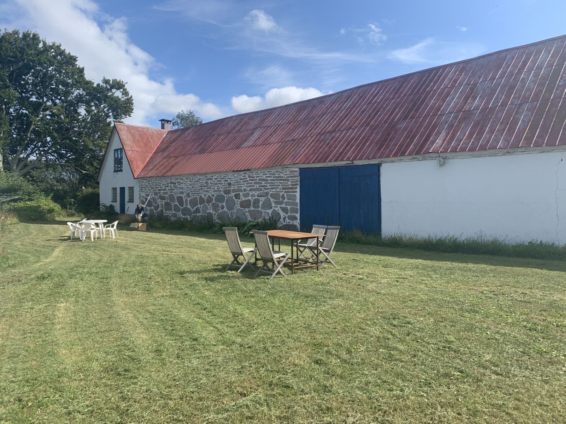 A stone building with a red roof, a white wall, and a blue door. There is a table and chairs in the foreground.
