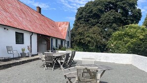 An outdoor seating area with tables and chairs, a white building with a red roof, and a large tree in the background.
