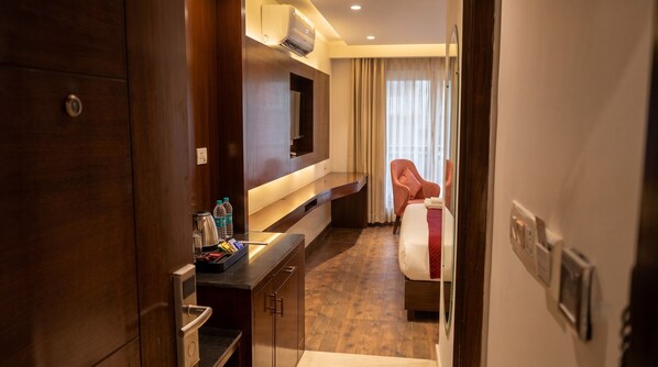 A hotel room with a wooden desk, a flat-screen TV mounted on the wall, a bed with a red blanket, and a window with curtains.