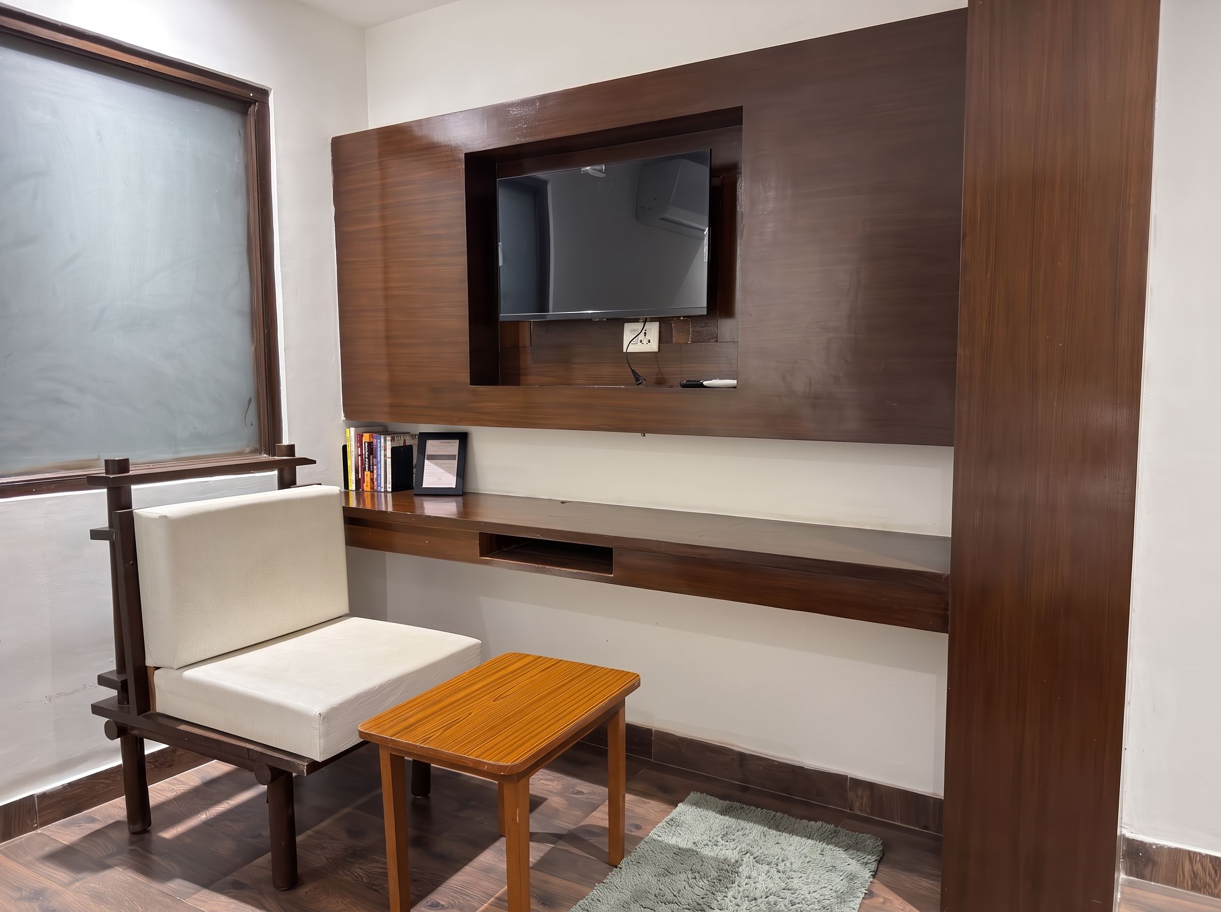 Una habitación con televisión integrada, una silla blanca acolchada, una pequeña mesa de madera y una alfombra gris.