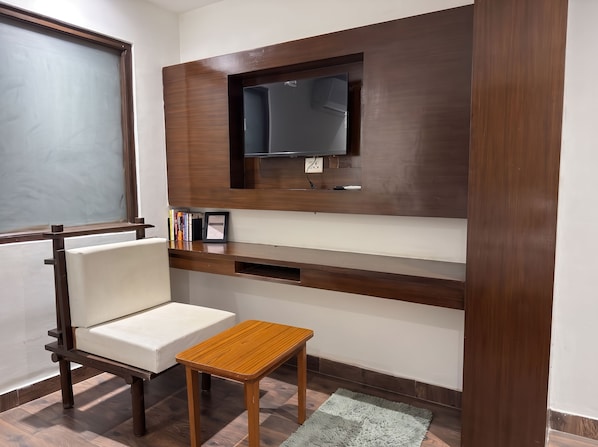 A room with a built-in TV, a white cushioned chair, a small wooden table, and a grey rug.