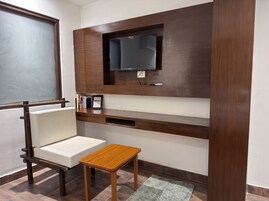 Una habitación con televisión integrada, una silla blanca acolchada, una pequeña mesa de madera y una alfombra gris.