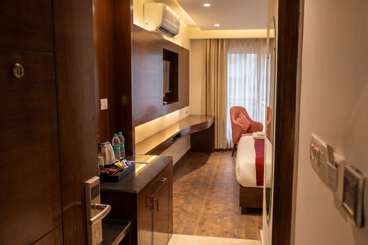 Habitación de hotel con un escritorio de madera, un televisor de pantalla plana montado en la pared, una cama con una colcha roja y una ventana con cortinas.