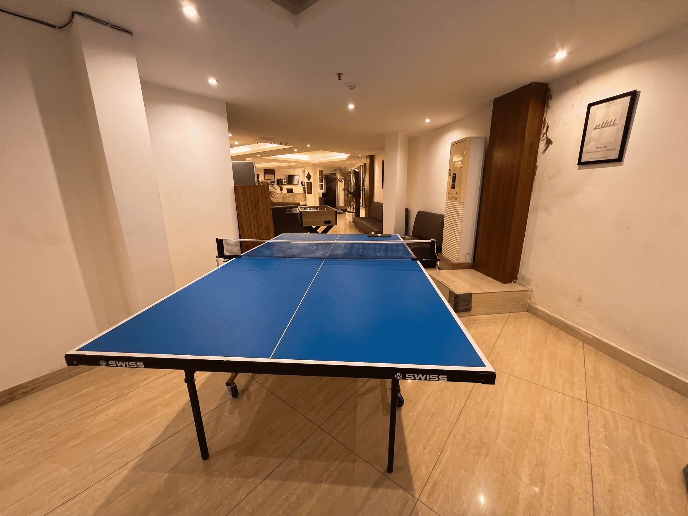Ein Tischtennistisch mit blauer Spielfläche und schwarzen Beinen, platziert in einem Raum mit Holzboden und weißen Wänden.