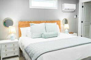 A neatly made bed with a white duvet and pillows, flanked by two bedside tables with lamps, a wall-mounted air conditioner, and a door. - Fall Special! Newly updated large home with pool! (St Simons Island)
