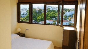 Quarto com cama, mesa de cabeceira, abajur, cômoda e janela com vista para casas e árvores.