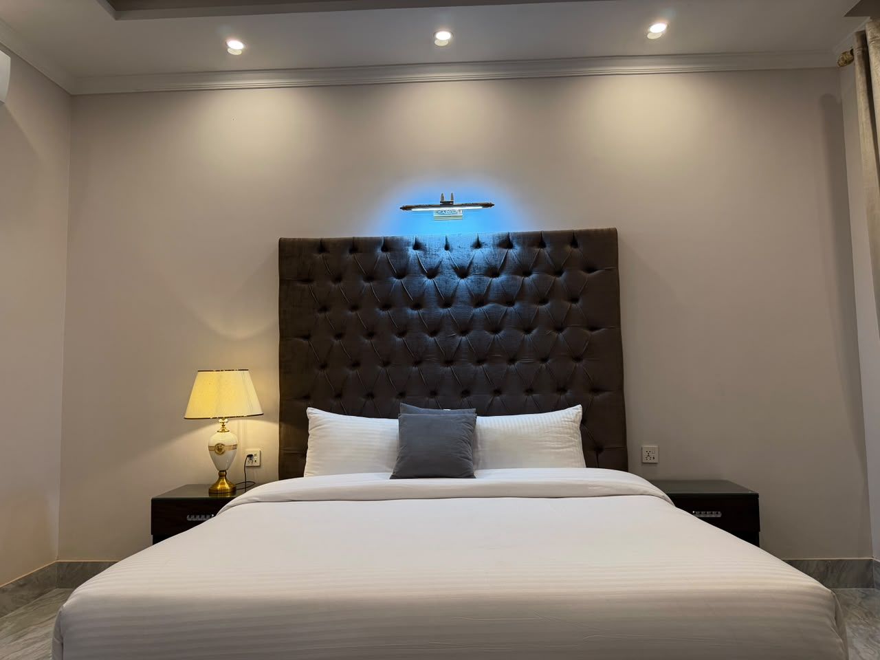 A hotel room with a large bed, a dark tufted headboard, a bedside lamp, and a wall-mounted light fixture.