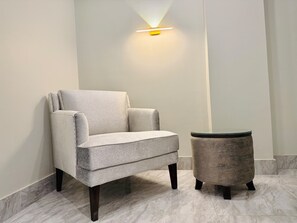 A modern room with a grey armchair and a round side table, both with dark legs, against a white wall with a wall-mounted light fixture. - Regency Suite Hotel (lahore)