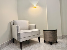 Una habitación moderna con un sillón gris y una mesita redonda, ambos con patas oscuras, frente a una pared blanca con una lámpara de pared.