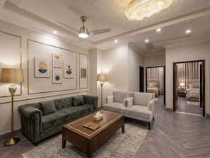 A modern living room with a green sofa, a white armchair, and a wooden coffee table. There are framed artworks on the wall, a ceiling fan, and a chandelier.
