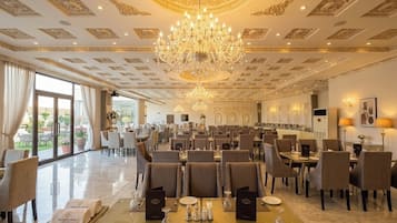 Uma ampla sala de jantar com várias mesas, elegantes lustres e vista para o exterior.