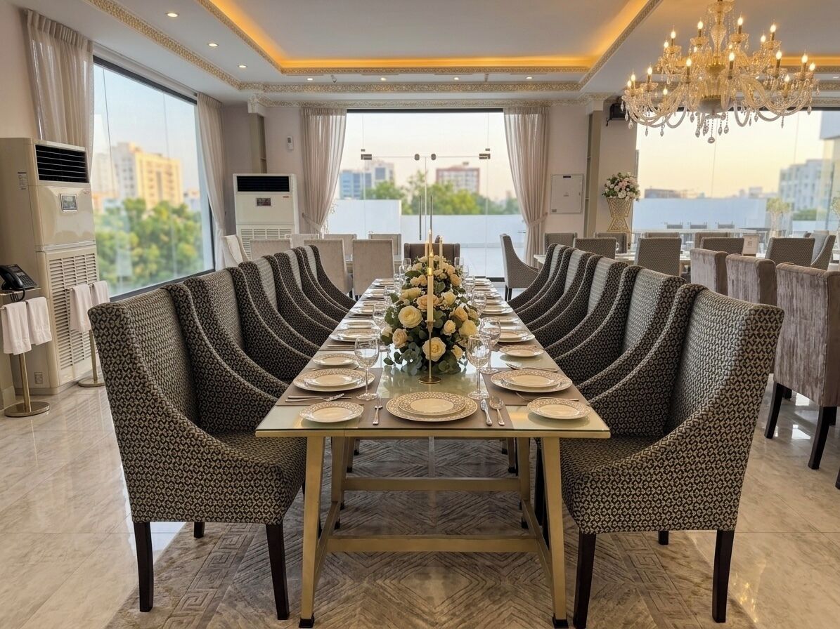 Comedor con una larga mesa dispuesta para una comida, rodeada de sillas tapizadas, un candelabro encima y vistas a la ciudad a través de amplias ventanas.