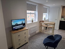 Una habitación con televisión mostrando noticias, un armario de madera, una pequeña mesa con sillas y una ventana con vista a un edificio de ladrillo.