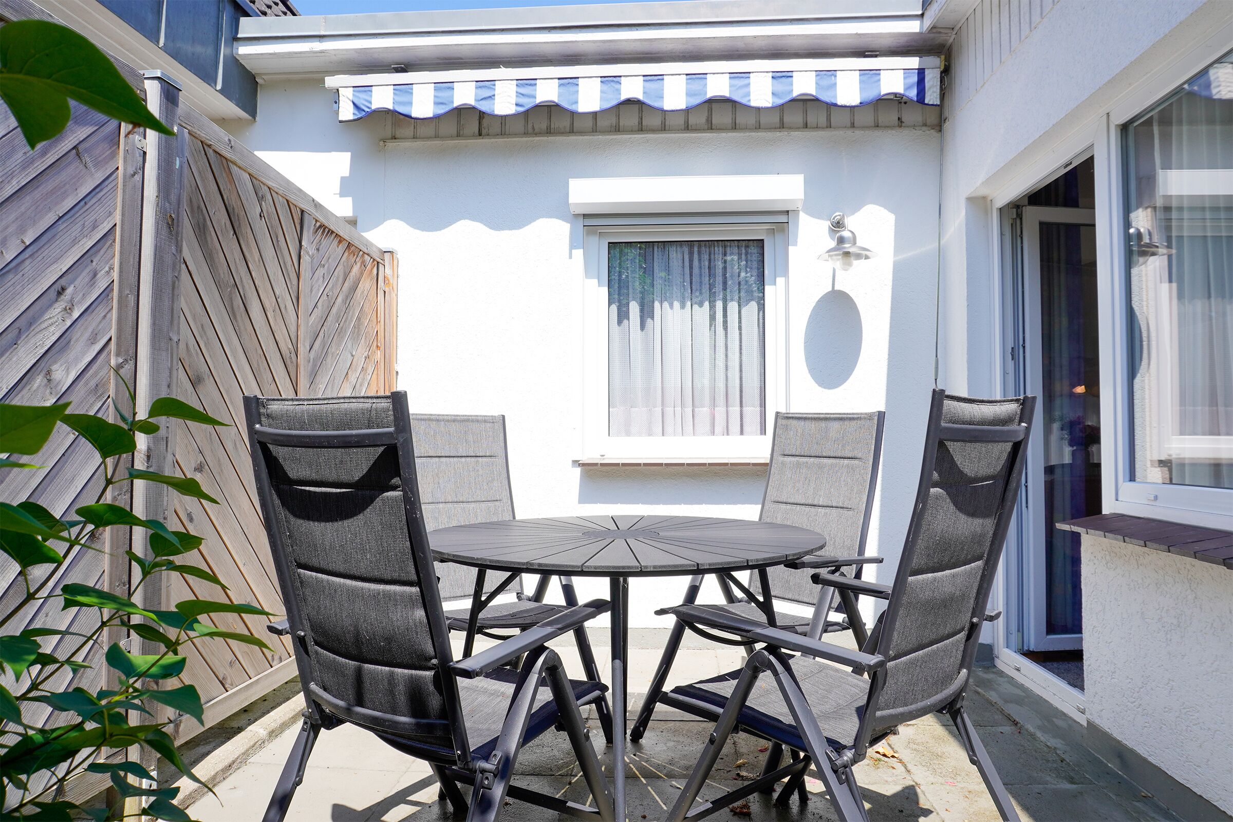 Eine Terrasse mit rundem Tisch und vier Stühlen, ein Holzzaun und ein Fenster mit Vorhang.