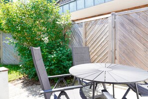 Eine Terrasse mit einem runden Tisch und zwei Stühlen, einem Holzzaun und einem grünen Busch.