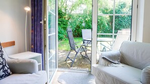 Ein Zimmer mit einer Couch, einem Sessel, einem kleinen Tisch und Blick durch eine Glastür auf einen Außenbereich mit Tisch und Stühlen.