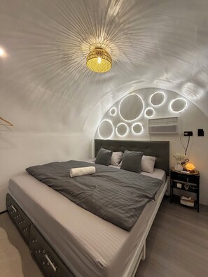 A modern bedroom with a curved ceiling, a bed with grey bedding, a bedside table with a lamp, and a wall-mounted air conditioner.