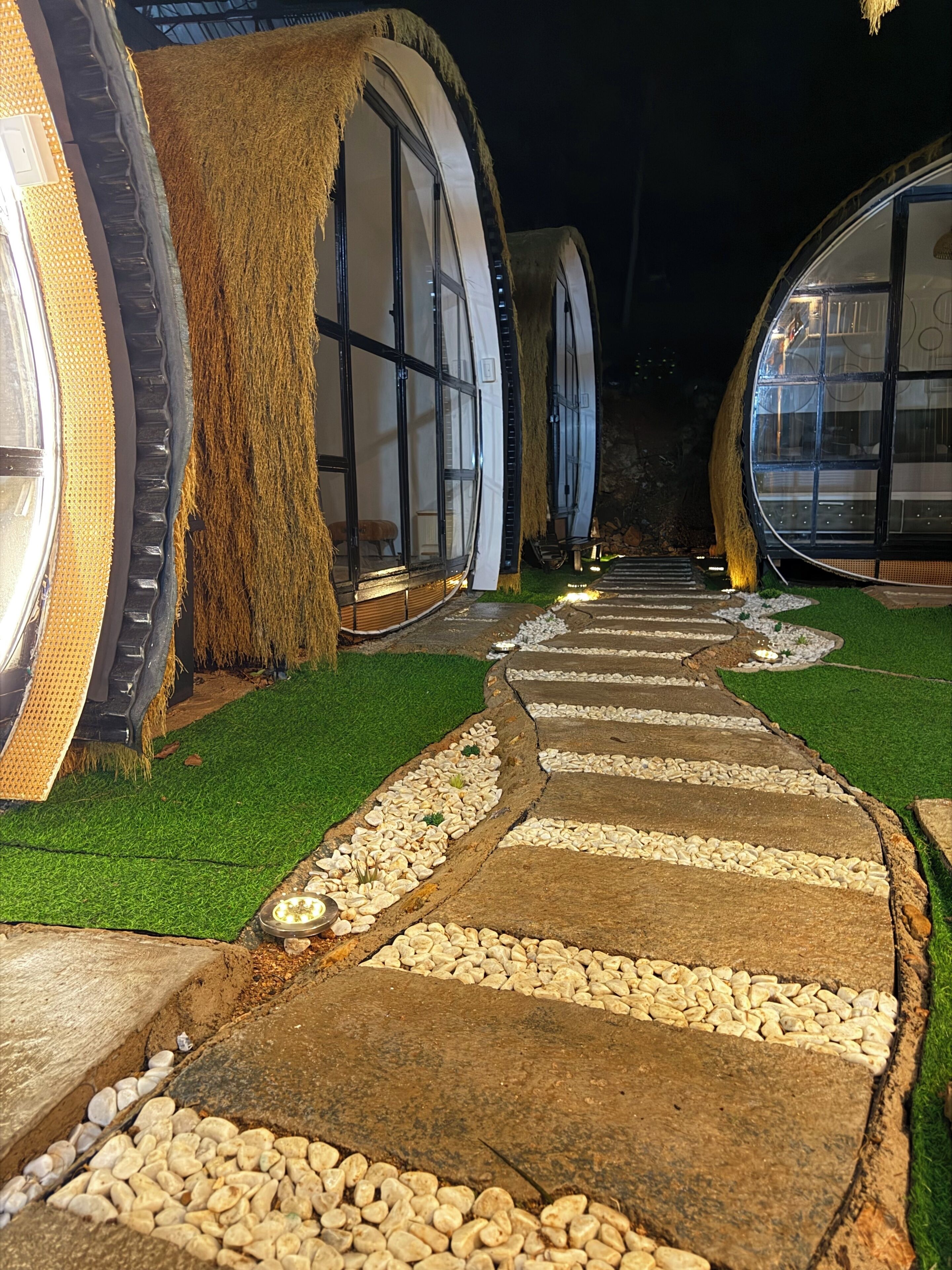 A pathway leading to circular structures with windows, surrounded by artificial grass and pebbles.