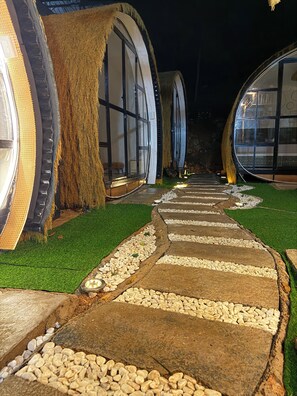 A pathway leading to circular structures with windows, surrounded by artificial grass and pebbles.