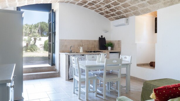 Una cocina con una mesa de comedor y sillas, una puerta abierta que da al exterior, y un pequeño rincón de descanso con un cojín.