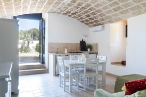 Uma cozinha com mesa e cadeiras, uma porta aberta para o exterior e uma pequena área de estar com um almofada.