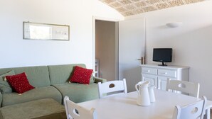 Una habitación con un sofá verde, una mesa blanca y sillas, un televisor sobre un mueble, y una puerta que da a otra habitación.