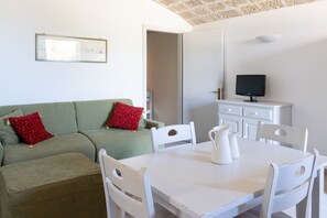 Ein Zimmer mit einem grünen Sofa, einem weißen Tisch und Stühlen, einem Fernseher auf einem Schrank sowie einer Tür zu einem anderen Raum.